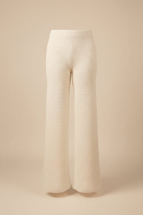 White crochet knit wide-leg pants displayed on a dress form mannequin in an indoor room with a wood floor and beige wall.