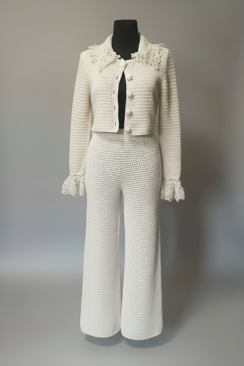 An off-white crochet knit three-piece suit displayed on a black mannequin against a neutral gray background. The set includes a cropped button-front jacket with a large decorative lace collar and ruffled cuffs, matching wide-leg trousers, and a coordinating top with a lace collar.