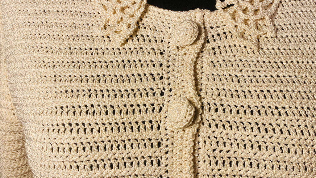 Close-up of a cream-colored crocheted cardigan sweater showing the textured knit pattern, decorative collar, and round front buttons.