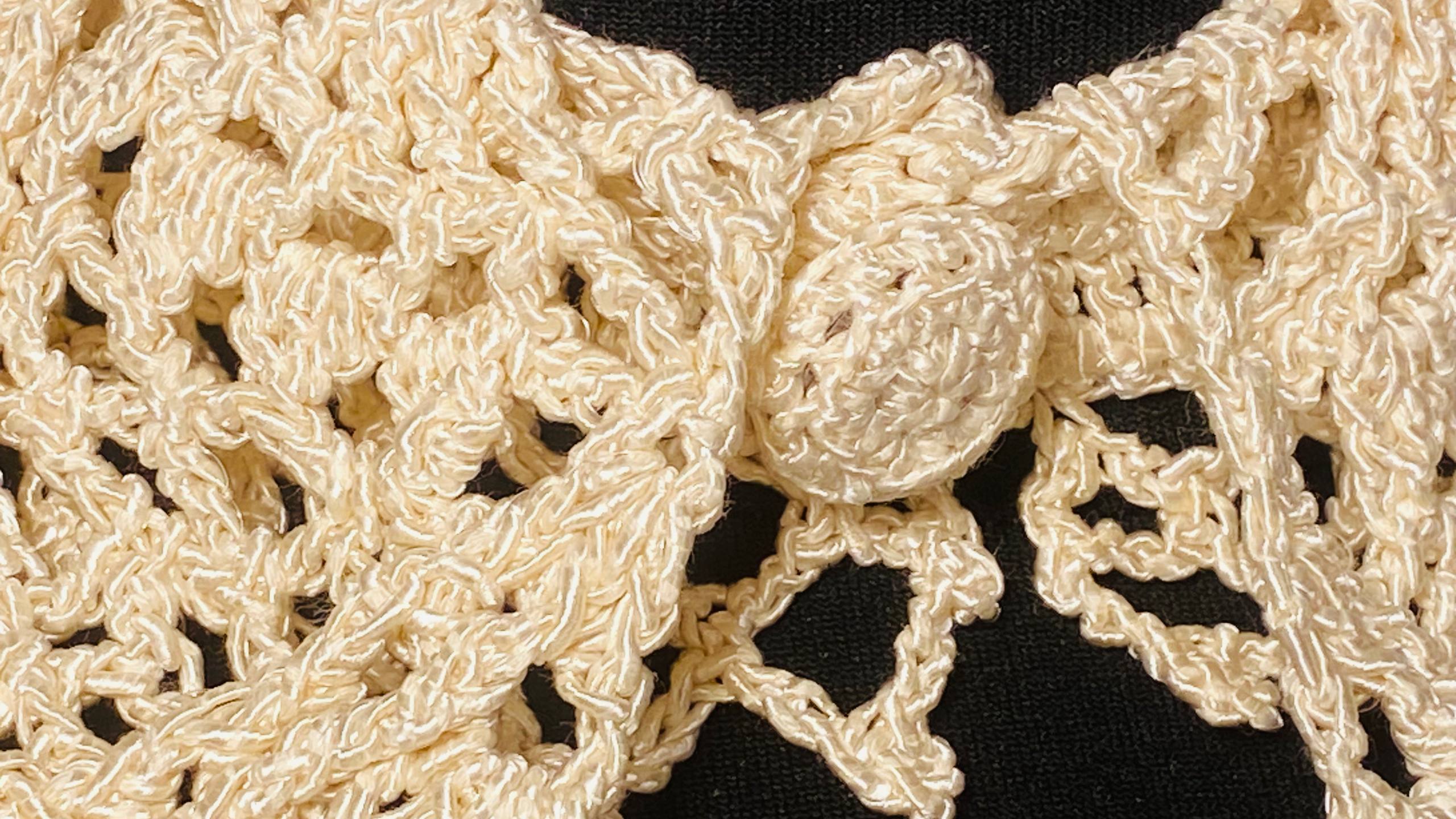 Close-up of a cream-colored crocheted lace item, likely a hat or garment trim, featuring a textured knit pattern and a decorative central loop, set against a black background.