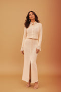 A woman models a matching two-piece knit set consisting of a cropped long-sleeve cardigan and a high-slit maxi skirt in a textured cream color, set against a solid warm tan studio background.