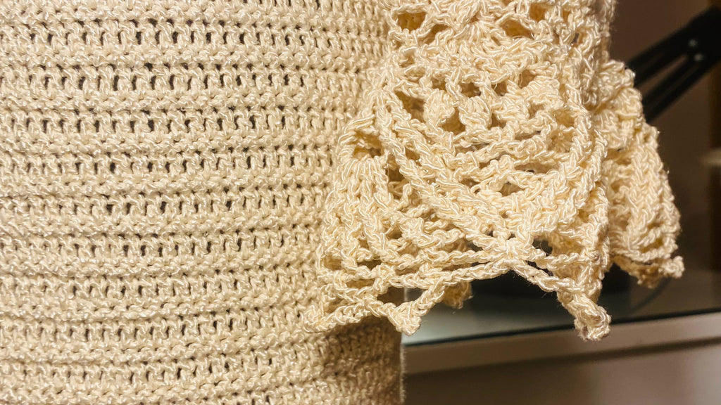 Close-up of a cream-colored crocheted textile, likely a blanket or throw, showing a textured knit pattern and an ornate lace-like border.
