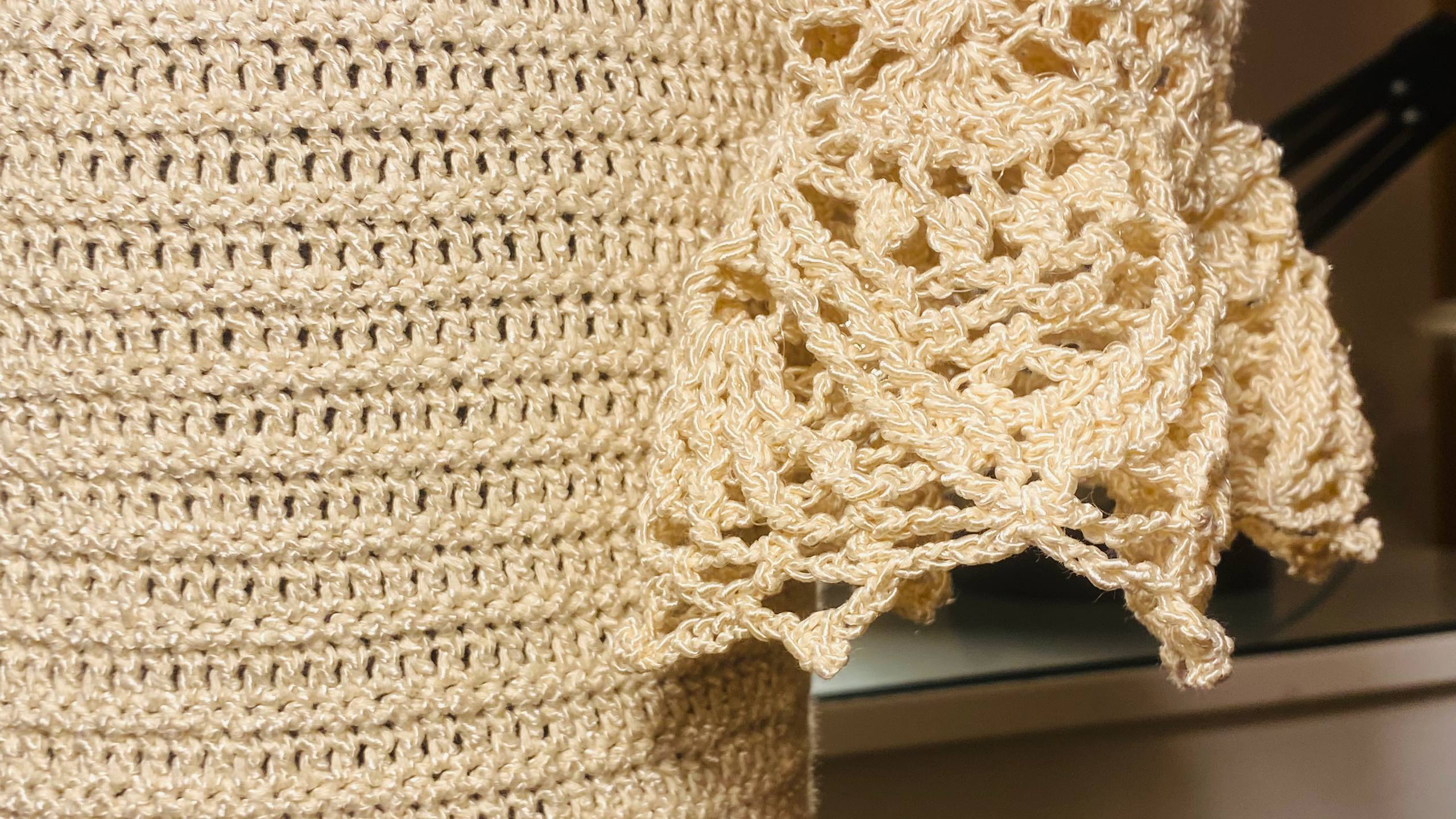 Close-up of a cream-colored crocheted textile, likely a blanket or throw, showing a textured knit pattern and an ornate lace-like border.