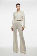 A model stands against a plain light gray studio background wearing a matching cream-colored knit two-piece set consisting of a cropped long-sleeve cardigan with a large ruffled collar and cuffs, and high-waisted flared trousers.
