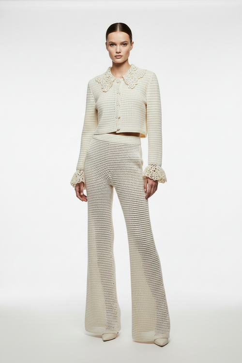 A model stands against a plain light gray studio background wearing a matching cream-colored knit two-piece set consisting of a cropped long-sleeve cardigan with a large ruffled collar and cuffs, and high-waisted flared trousers.