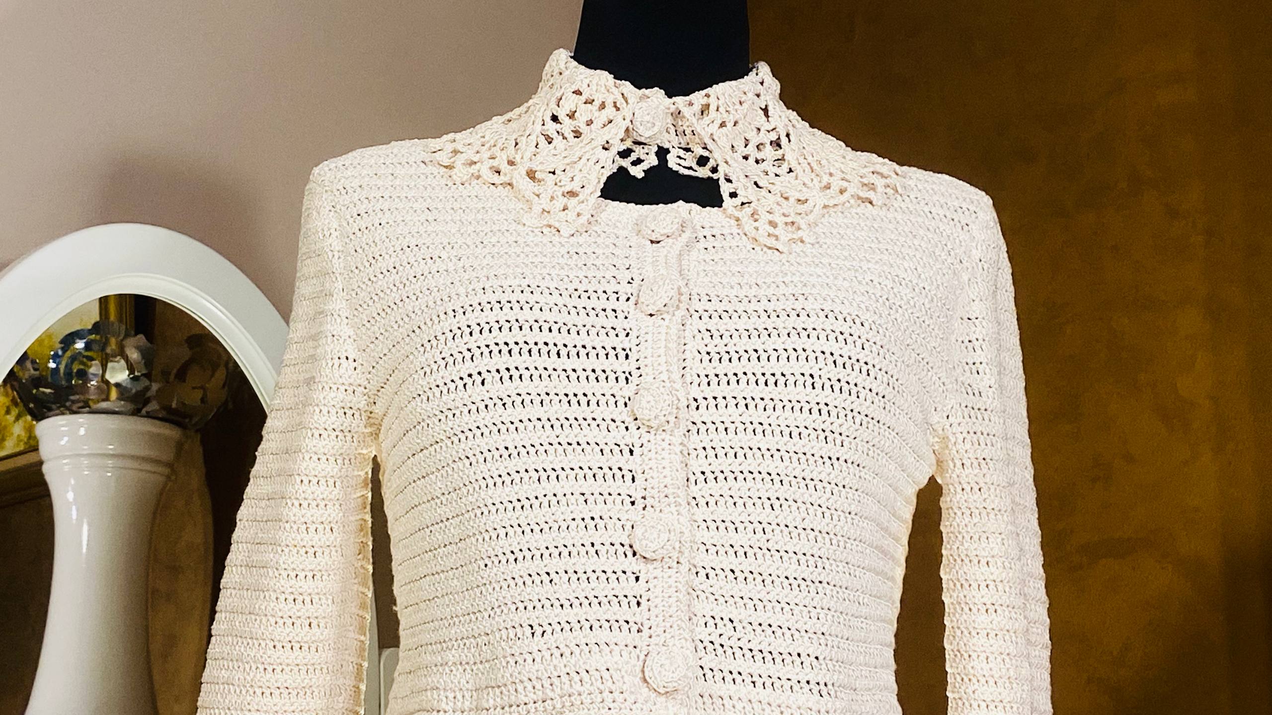 An off-white crocheted cardigan sweater with a lace collar and button-front closure, displayed on a black mannequin torso against a warm brown background.