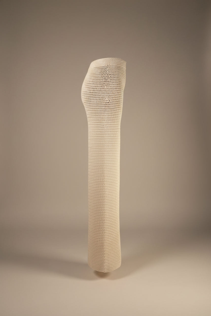 A tall, cylindrical white knit or crocheted support sleeve standing on a stand in an indoor room with wood flooring and a potted plant in the background.