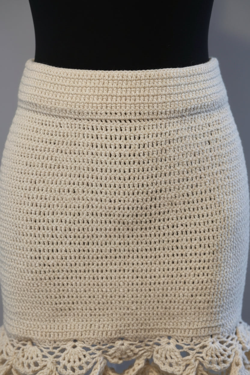 A close-up of a cream-colored crochet mini skirt displayed on a black mannequin torso against a neutral gray background.