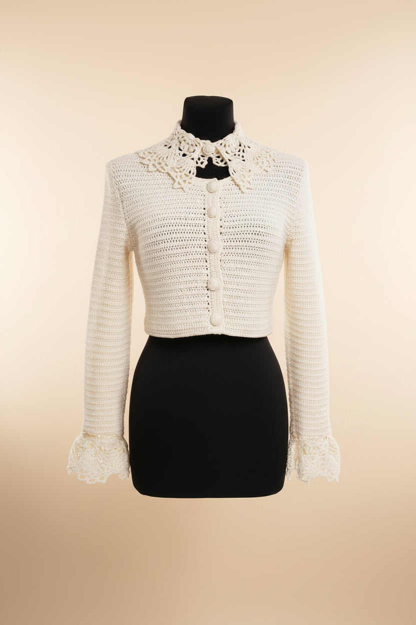 An off-white cropped knit cardigan with a large floral lace collar and matching lace cuffs, displayed on a black dress form mannequin against a warm beige background.